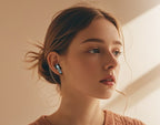 A close-up lifestyle shot of a woman with light brown hair in a bun, wearing a small silver and black in-ear wireless earbud. She is wearing a textured terracotta knit top against a warm beige background with soft diagonal light rays.