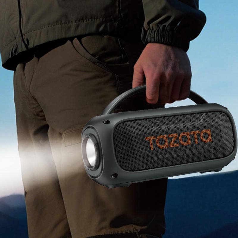 A person outdoors carrying a dark gray portable Bluetooth speaker with a built-in handle and a visible front light. The speaker has the brand name 'TAZATA' printed in orange on the front grille.