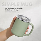 A hand holds a matte sage green insulated mug with a clear lid and silver rim. The background is a light gray textured wall with text overlays reading 'SIMPLE MUG' and 'Iced Americano for hot days / Hot Latte for cold days'.